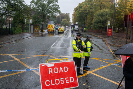 A police cordon surrounds a synagogue in London. Emergency vehicles are visible in the background. The area is quiet, suggesting the immediate aftermath of an incident. The image emphasizes security and investigation.