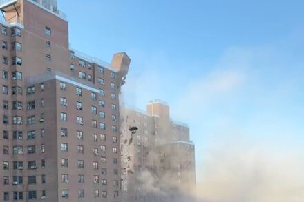 Debris falls from the side of a tall, grey building in the Bronx, scattering across the street below. A person in a hard hat inspects the scene.