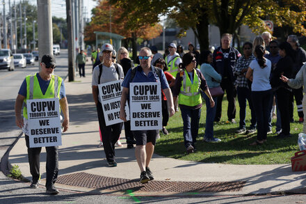 A line of Canada Post workers on strike, holding picket signs outside a post office.