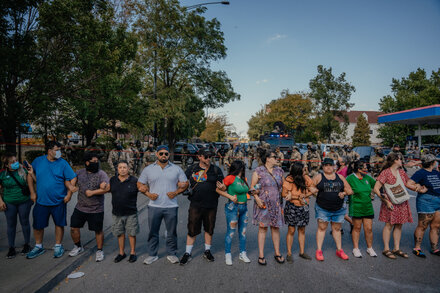 A group of diverse people gathered on a street in a residential Chicago neighborhood, some holding signs, looking concerned or engaged in conversation. The scene suggests a community event or gathering related to social issues.