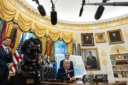 Donald Trump speaks at a podium with the White House in the background, addressing a crowd of people. Police and protestors are visible in the distance.