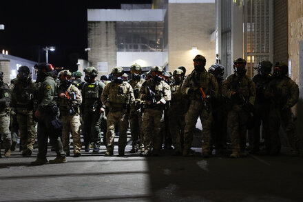 Federal agents in riot gear stand guard outside a federal courthouse in Portland, Oregon, with smoke in the background.