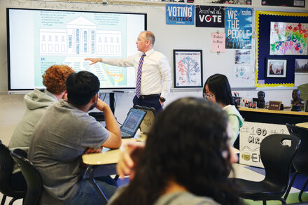 A diverse group of high school students actively engaged in a classroom discussion, raising hands and speaking with a teacher near a whiteboard, symbolizing the challenges and importance of civics education.
