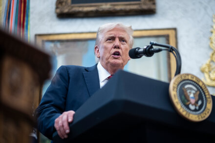 James Comey testifying in court with a defiant expression, while a blurred image of Donald Trump is visible in the background, possibly on a screen or a banner, suggesting a contentious legal battle.