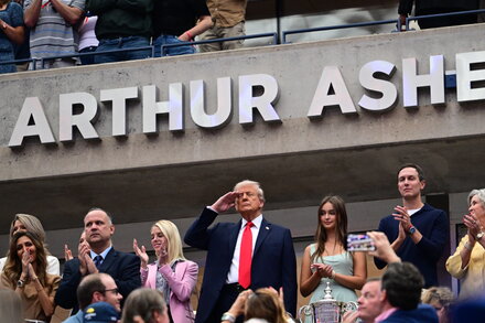 Donald Trump in a suit, clapping and smiling at a sporting event, possibly a baseball game, with a crowd in the background.