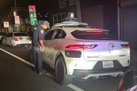 A Waymo self-driving car making an illegal U-turn on a city street, with a police officer observing the incident.