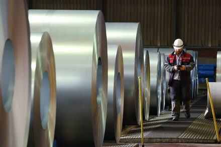 A close-up shot of stacks of finished steel sheets in a factory, glinting under industrial lights. The textured surface of the steel shows subtle variations in color, from dark gray to metallic silver. In the background, out of focus, more machinery and perhaps workers can be vaguely perceived, suggesting a bustling industrial environment.