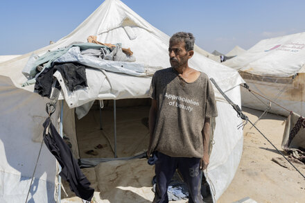 A family huddles together outside a makeshift tent in a barren landscape, with debris scattered around. The parents look tired, and children are partially visible. The sky is overcast.