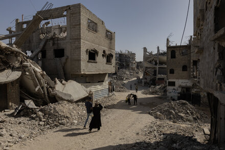 Smoke rises from buildings in a Gaza neighborhood after an Israeli airstrike, with debris scattered in the foreground. People are seen looking at the damage.