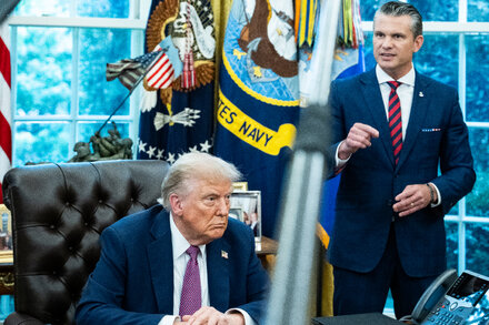 Pete Hegseth and Donald Trump stand side-by-side, both in dark suits, facing a group of uniformed military officials in a formal setting. Trump appears to be addressing the officials while Hegseth listens attentively.