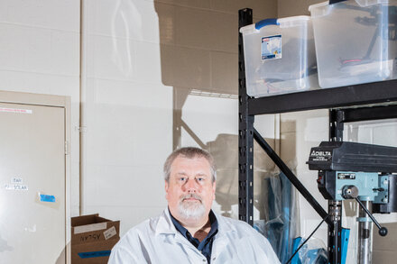 Portrait of a scientist in a lab coat smiling next to a complex piece of spacecraft equipment, a feature image for an article about his lab testing cutting-edge spacecraft.