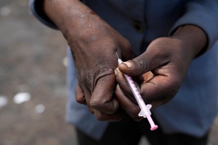 A close-up of a person's arm, showing a needle inserted into a vein. The lighting is dim, suggesting a clandestine or unhealthy setting. The skin around the injection site appears bruised or scarred.