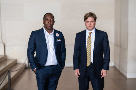 Two lawyers stand in a hallway, looking serious. One is a woman with short dark hair, wearing a suit jacket and holding a document. The other is a man with glasses, wearing a suit. They appear to be in a government building.
