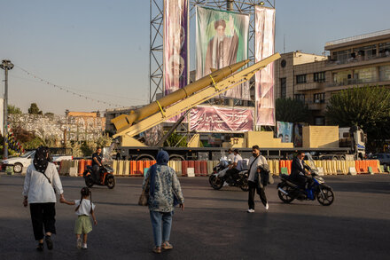 Iranian flags flying near a building, with a crowd of people in the foreground. The image represents Iran's role in the Israel-Hamas conflict.