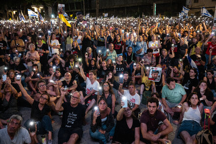 An aerial view of a large protest in Israel, with thousands of people gathered in a public square holding signs and Israeli flags.