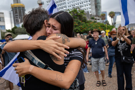 An Israeli woman smiles while looking at her phone, with other people in the background, likely reacting to news of a deal or ceasefire.
