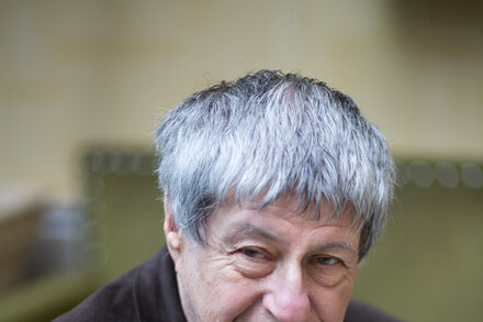 Ivan Klima, a Czech novelist, sits at a table, looking thoughtful. He has short, gray hair and is wearing a collared shirt. The background is a simple, blurred indoor setting.