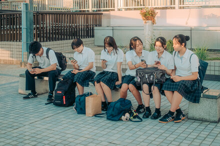 A group of people walking through a bustling Japanese city street, with many of them looking at their smartphones. In the foreground, one person is smiling and looking up, not at a device.