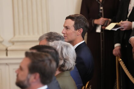 Jared Kushner, former senior advisor to the president, stands in a formal setting, possibly a conference room or a government building. He is wearing a suit and tie, looking directly at the camera with a neutral expression. The background is slightly blurred but suggests an interior space with warm lighting.