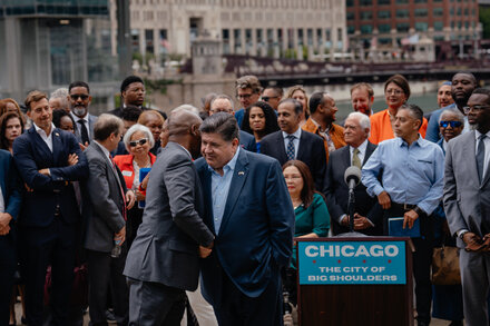 Illinois Governor JB Pritzker speaking at a podium with microphones, gesturing with his hand during a press conference.