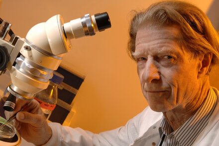 John B. Gurdon, a distinguished scientist with white hair and glasses, smiles slightly while wearing a dark suit and tie, sitting in a lab-like setting with scientific equipment blurred in the background.