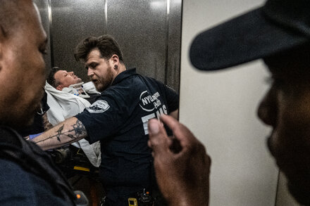 A journalist with a camera is caught in a chaotic scene outside a courthouse, surrounded by various people and security personnel.