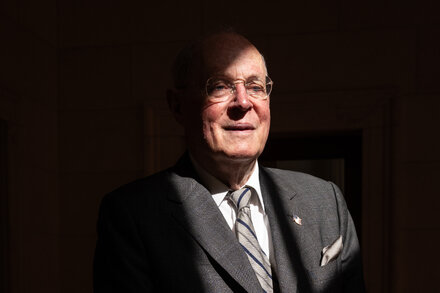 Justice Anthony Kennedy speaks at an event, gesturing with one hand. He is wearing a dark suit, light shirt, and patterned tie. A microphone stand is in front of him.