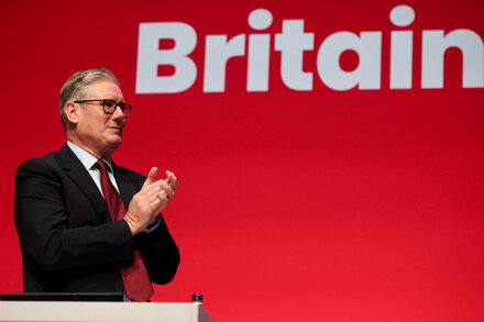 Keir Starmer, leader of the Labour Party, speaks at a podium in front of a crowd, with the Labour Party logo visible. He appears serious and determined.