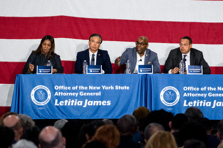 New York Attorney General Letitia James stands at a podium, with a backdrop displaying the New York State seal, speaking to a room full of microphones.