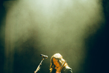 Lucy Dacus, dressed in a black suit, stands on a dimly lit stage holding a microphone, officiating a wedding for a couple. The couple, a man and a woman, are smiling and holding hands in front of her. Concert lighting creates a warm atmosphere.