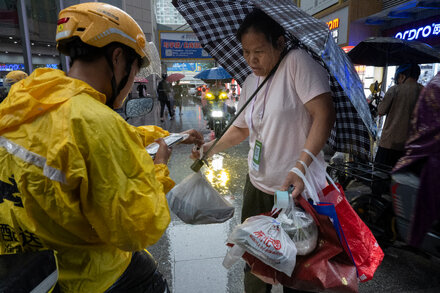Delivery rider on a motorcycle carrying a large food delivery bag, navigating a busy city street with tall buildings in the background, implying last-mile delivery to high floors.