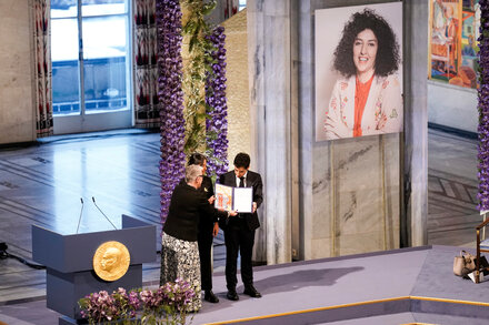 Maria Corina Machado, a prominent Venezuelan opposition figure, stands with a serious expression. She is wearing a dark suit jacket and a white blouse, looking directly at the camera. The background is slightly blurred, suggesting an indoor setting.