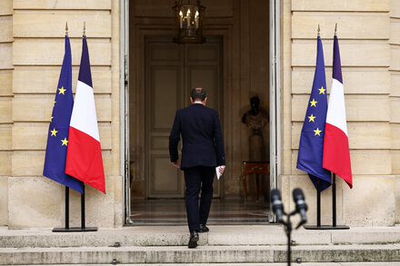 French President Emmanuel Macron stands in a formal setting, looking serious, with a blurred background suggesting a government building or official event. The image captures him in a moment of reflection or address, conveying the weight of his political situation.