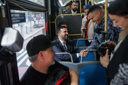 Assemblyman Zohran Mamdani riding a crowded M60 Select Bus in New York City, discussing his plan to improve bus speeds.