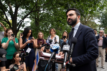 Assemblyman Mamdani speaks into a microphone at a podium, with a banner behind him that reads 'New York State Assembly'. He is looking intently at the audience.