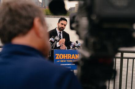 Representative Zohran Mamdani speaking at an outdoor rally in a suburban setting, surrounded by diverse supporters.