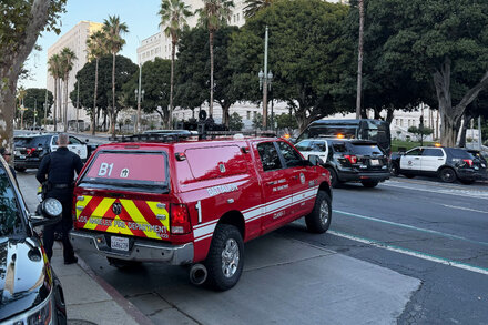 Police officers surround a white sedan stopped on a sidewalk in front of Los Angeles City Hall, with a man being escorted by police nearby.