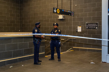 A police officer stands near a subway station entrance in New York City. The scene appears to be an active investigation.