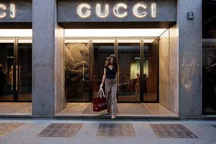 A woman confidently walks through a high-end fashion boutique, surrounded by luxurious clothing racks and modern decor. She is looking towards the camera with a slight smile, holding a designer shopping bag. The store appears spacious and well-lit, emphasizing the elegant display of apparel.