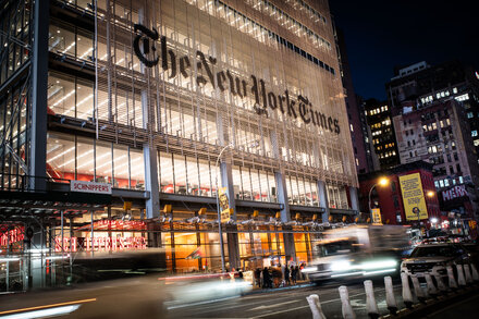 Three individuals in handcuffs being escorted by police officers in front of the New York Times building, which shows signs of graffiti and spray paint on its lower facade.