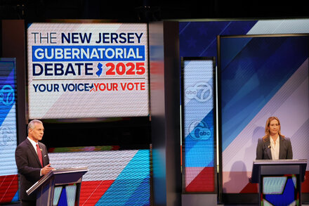 Two male candidates stand at podiums on a debate stage, facing an audience. One wears a dark suit, the other a lighter suit. A large screen behind them displays the debate topic. The atmosphere is serious and focused.