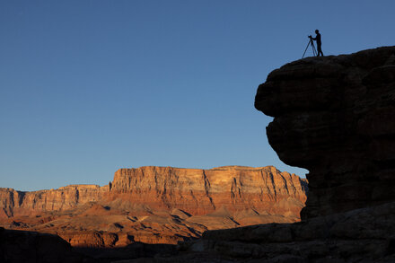 A lone ranger stands at the entrance of a national park, with a 'Closed' sign prominently displayed. The sky is overcast, and the surrounding landscape shows dormant trees, hinting at autumn or winter. The image conveys the impact of a government shutdown on public access to national parks.