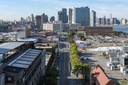 An aerial view of the New York City skyline, focusing on a dense residential area with numerous tall buildings and construction sites near a waterfront. The sky is clear, and the overall impression is one of urban development and expansion.