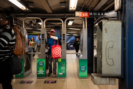 A New York City subway car pulls into a station platform, seen through the window of another train, with 'MTA' logo visible.