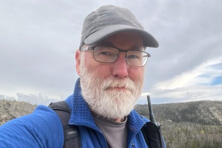 Man in a rustic cabin, looking surprised while holding a satellite phone or device, with a remote, natural landscape visible outside a window.
