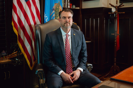 Oklahoma Governor Kevin Stitt stands at a podium, with an American flag visible in the background, speaking to a crowd. He is wearing a suit and tie, looking serious. The image captures him mid-speech, conveying his address on a significant topic.