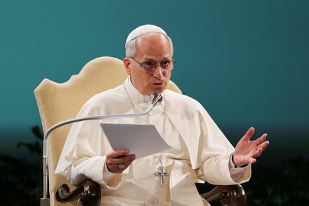 Pope Leo stands at a podium in a grand hall, addressing an audience. He wears white papal vestments and a skullcap, his hands gesturing as he speaks. The hall features classical architecture with ornate columns and high ceilings, and a large window behind him lets in light. The atmosphere is formal and solemn.