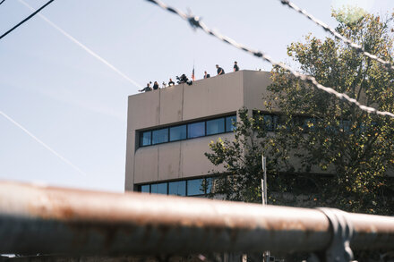 A group of right-wing influencers and activists gathered in Portland, Oregon, near a protest, with a blurred cityscape in the background. Some individuals are holding flags or signs, while others are engaging in discussions.