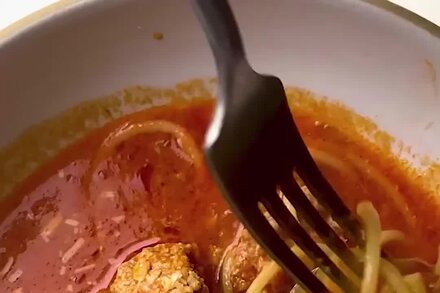 A bowl of spaghetti and chicken meatball soup with herbs, served with a spoon.