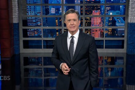 Stephen Colbert looking serious, with a microphone in front of him, against a dark blue background, from his late-night show